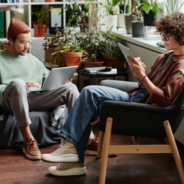 Two young serious coworkers using wireless mobile gadgets while sitting in armchairs in openspace office with variety og green plants